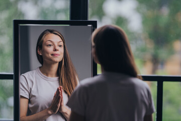 Reflective young woman in a casual t-shirt practicing self-affirmation in front of a mirror in a...