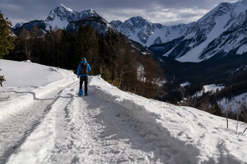 Fototapeta na wymiar Man in snow shoes on hiking trail with panoramic view on snow capped mountain peaks of Karawanks, Carinthia, Austria. Looking at summits Kosiak and Hochstuhl, Austrian Alps. Alpine landscape in winter