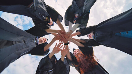 Team of college or university students celebrating graduation. Group of happy successful graduates in academic hats and robes standing in circle and putting their hands together.