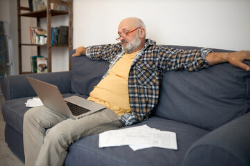Side view of perplexed tired male pensioner sitting in couch with laptop on knees and outstretched...