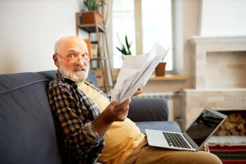 Side view of worried anxious elderly man in glasses looking at camera with panic facial expression holding overdue bills, trying to do online payment using laptop with copy space screen