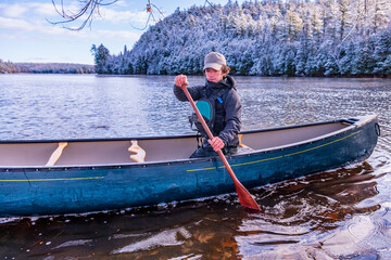 young man paddling a green canoe solo on a river with trees clad in freshly fallen snow in the...