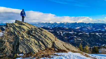 Man with backpack standing on massive rock formation at Steinerne Hochzeit, Saualpe, Lavanttal Alps, border Styria Carinthia, Austria, Europe. Panorama of alpine meadows and snowcapped mountains