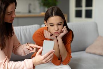 Angry young mother showing cellphone to upset teenage girl while daughter looking at screen with indifference, sitting together on sofa at home - obrazy, fototapety, plakaty