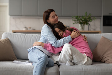 Loving mother hugging teen daughter, mom showing understanding and support to upset sad teenage girl, sitting together on couch at home