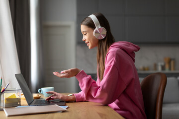 Teen schoolgirl in headphones studying online with remote tutor from home, student using laptop...