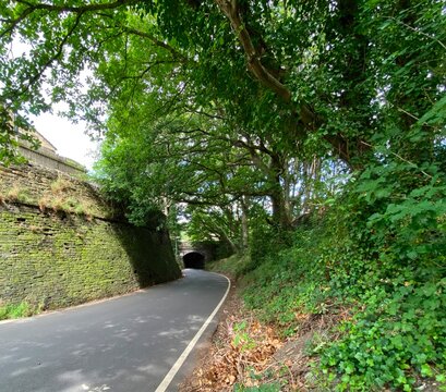 Wood Lane, curving toward a railway bridge, with a high stone wall, trees, and a cloudy sky in, Colnebridge, UK