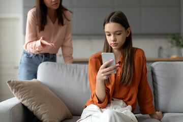 Angry mother shouting and arguing with teenage daughter over use of mobile phone, girl sitting on sofa at home, ignoring mom