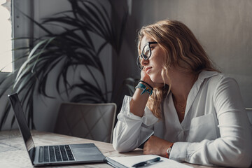 Discontented thoughtful woman with hand under chin bored at work, looking away sitting near laptop,...