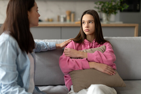 Mother consoling her depressed teen daughter, sitting on sofa at home, girl embracing pillows. Teenager problems