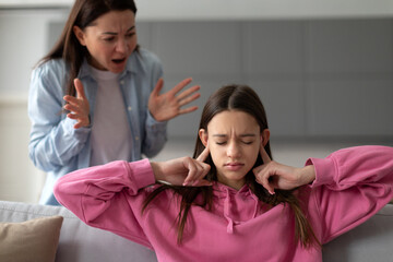 Strict mother scolding teenage daughter, mom lecturing teen girl, child covering ears with fingers, sitting on sofa. Parental harsh discipline, adolescent problem behavior