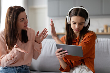 Teenage girl in headphones using digital tablet and gesturing stop to mother, while mom scolding daughter, sitting on sofa at home - obrazy, fototapety, plakaty
