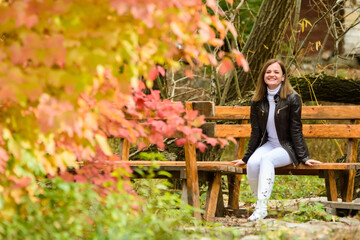 A young beautiful girl sits on a bench in an autumn park and looks happily into the frame