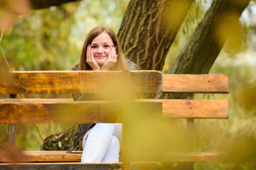 A young beautiful girl sits on a bench in an autumn park and looks cheerfully into the frame