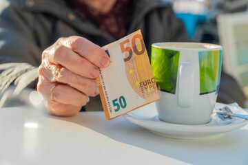  Hands of an older woman with a 50 euro bill next to a cup of coffee.