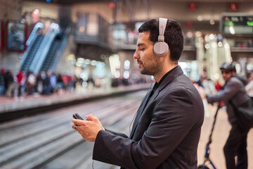 latin man waiting for the train while using his smart phone with headphones on playing games or...