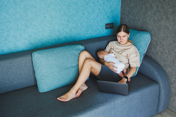 A smiling young mother is breastfeeding her baby and working at home with her baby on the sofa...