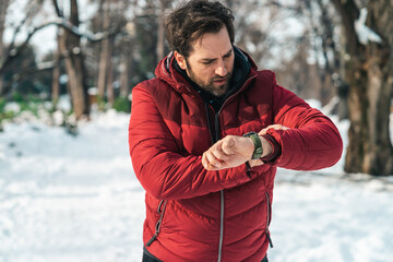 Serious mid adult man in red winter jacket looking at his wrist watch while taking a walk on snowy...