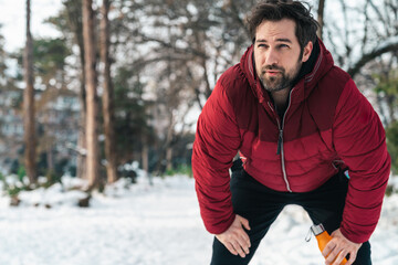 Serious handsome man taking a break from jogging, resting and holding water bottle while exercising in winter.