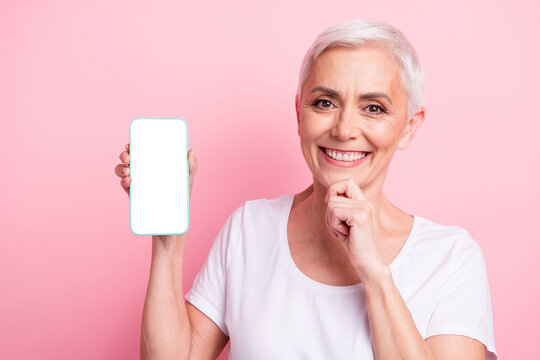 Photo of intelligent senior woman with short hairstyle wear white t-shirt touch chin show smartphone screen isolated on pink background