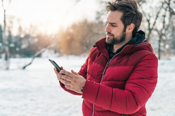 Handsome serious adult man using mobile phone while walking in public park on a winter morning,...