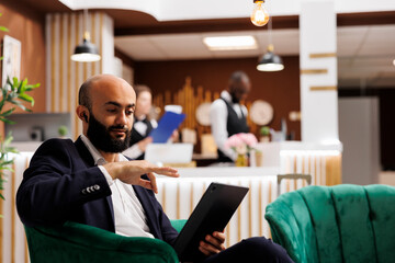 Middle eastern man on videocall using tablet, waiting to register in hotel lounge area. Employee...