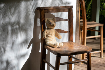 Old 1960's vintage Teddy bear sitting on a wooden chair on a porch outdoors with dramatic shadows.