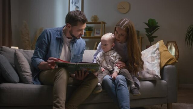 Little baby girl reading book with parents. Happy family sitting on couch and spending time together. Little boy looking at picture in book. Mother and father reading tales to little son.