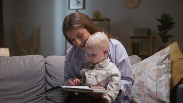 Mother spending time together with little son. Mother reading book to kiddo. Mother reading tales to toddler. Woman enjoying being mother. Little boy attentively looking at pictures in book.