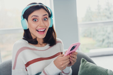 Portrait of astonished cheerful lady sit chair use smart phone listen music headphones pastime...