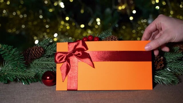 A hand places a orange gift certificate with a red satin ribbon and a bow on a brown tablecloth on a New Year's branch