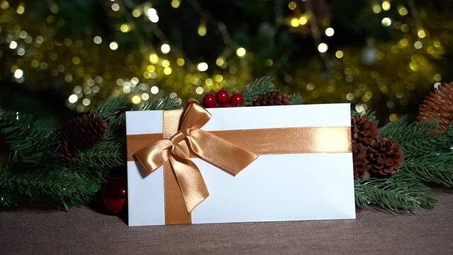 A hand places a  white gift certificate with a gold satin ribbon and a bow on a brown tablecloth on a New Year's branch