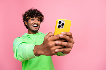 Photo of positive indian guy wear green hoodie laughing holding his smartphone using for promoting...