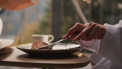 Lady uses knife and fork at breakfast in cottage. Woman has pancakes for morning meal by panoramic window with view on nature. Woman cuts pancakes