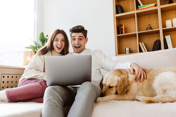 Young overjoyed couple using laptop computer shopping online sitting on comfortable sofa at home....