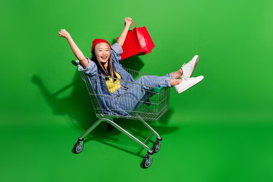 Full body photo of attractive young woman ride trolley win shopping bags dressed stylish denim clothes isolated on green color background