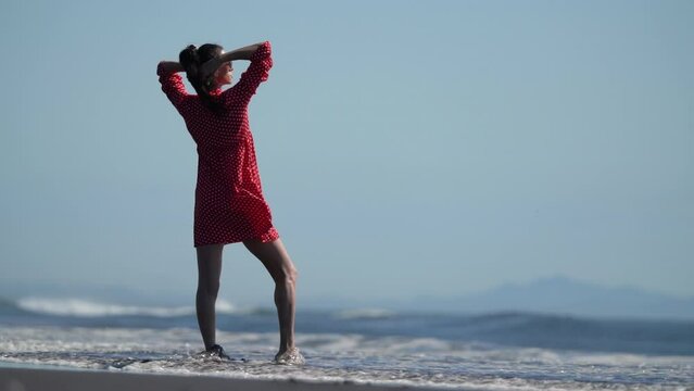 Serene woman standing ankle deep in water breaking seawave on sandy beach of ocean. Full length model generation x dressed in red polka dot babydoll dress loose fitting, hands behind head. Slow motion