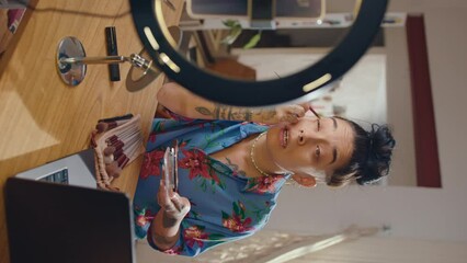 Young female beauty blogger sitting in front of a ring light with phone, doing eye makeup and telling some hacks on camera while making a social media video. Vertical format footage