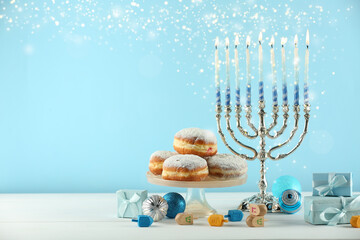 Hanukkah celebration. Menorah with burning candles, dreidels, gift boxes and donuts on white wooden...