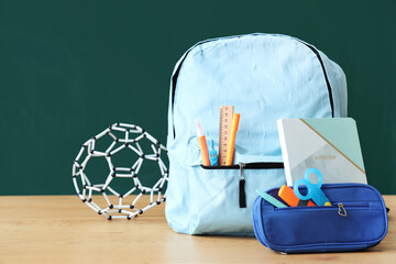 Backpack with school stationery and molecular model on table near green chalkboard