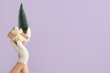 Female hands holding Christmas gift and decorative pine tree on lilac background