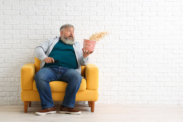 Shocked mature man with falling popcorn sitting in armchair near white brick wall