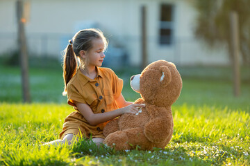 Pretty child girl tenderly embracing her teddy bear friend outdoors on green grass lawn. Concept of...