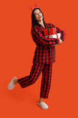 Beautiful young woman in checkered pajamas and reindeer horns with Christmas gift boxes on orange background