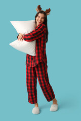 Beautiful young woman in checkered pajamas and reindeer horns with soft pillow on blue background