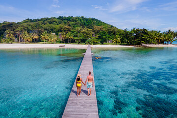 couple walking at a wooden pier in the ocean of Koh Kham Trat Thailand, aerial view of the tropical island near Koh Mak Thailand. white sandy beach with palm trees and big black boulder stones  - obrazy, fototapety, plakaty