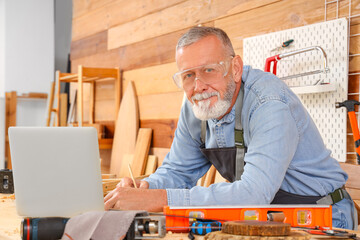 Mature carpenter working with laptop at table in shop