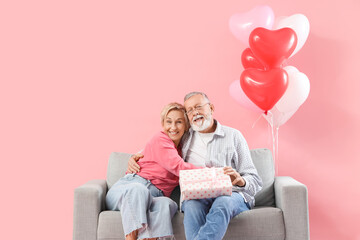 Mature couple with gift hugging on sofa against pink background. Valentine's Day celebration