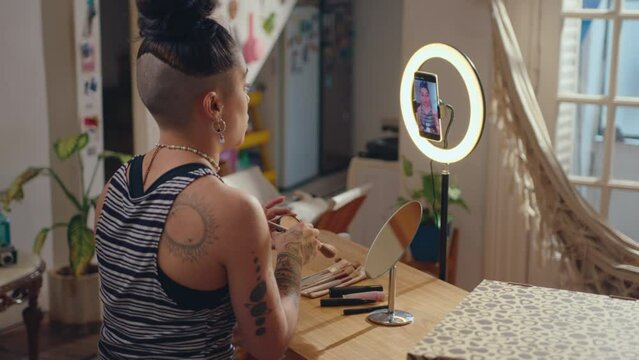 Female beauty blogger using phone with ring light for filming video tutorial or going live on social media. She applying bronzing powder with brush and telling makeup hacks on camera