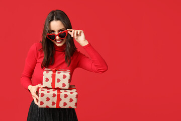 Beautiful young woman in stylish heart shaped sunglasses with gift boxes on red background....